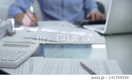 Calculator, pen and documents are lying on accountant's desk. Businessman wearing blue shirt is working with documents on the background. Business people, audit concept Calculator, pen and documents are lying on accountant's desk. Businessman wearing blue shirt is working with documents on the background. Business people, audit concept 132708540
