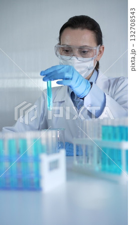 Female lab worker or scientist, wearing coat, blue medical gloves, mask and protective glasses is examining test tube with blue liquid in modern laboratory. Low key lighting photo 132708543