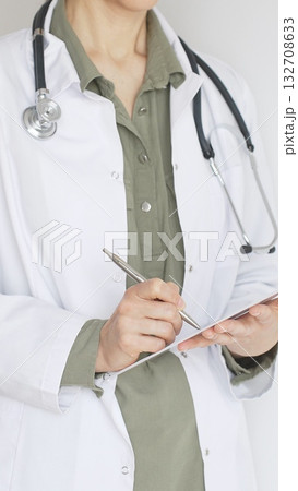 Female doctor, wearing a stethoscope and a medical coat over a green blouse, is standing and writing on a white clipboard, representing medical expertise and patient care. Medicine concept. Female doctor, wearing a stethoscope and a medical coat over a green blouse, is standing and writing on a white clipboard, representing medical expertise and patient care. Medicine concept. 132708633