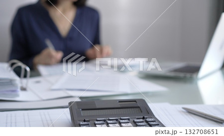 Black calculator, pen and documents are lying on accountant's desk. Businesswoman wearing blue dress is working with documents on the background. Business and audit concept Black calculator, pen and documents are lying on accountant's desk. Businesswoman wearing blue dress is working with documents on the background. Business and audit concept 132708651