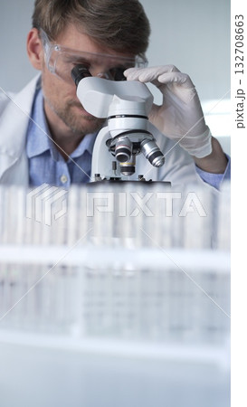 Man scientist wearing lab coat, white gloves and protective glasses using microscope doing research in modern laboratory, portrait view. Low key lighting photo. Science and Medicine 132708663