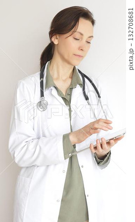 Doctor woman wearing white medical coat over green blouse, stethoscope, is reviewing patient records on digital tablet, standing in medical workspace with natural lighting. Medicine concept Doctor woman wearing white medical coat over green blouse, stethoscope, is reviewing patient records on digital tablet, standing in medical workspace with natural lighting. Medicine concept 132708681