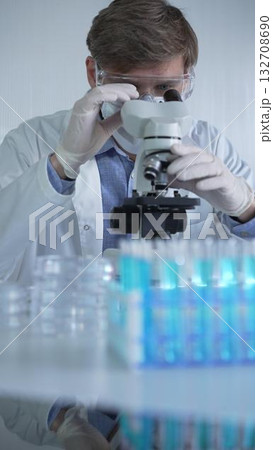 Male scientist researcher wearing lab coat, white protective gloves, medical mask and glasses, is using microscope in modern laboratory setup, portrait view. Low key lighting photo. Science Male scientist researcher wearing lab coat, white protective gloves, medical mask and glasses, is using microscope in modern laboratory setup, portrait view. Low key lighting photo. Science 132708690