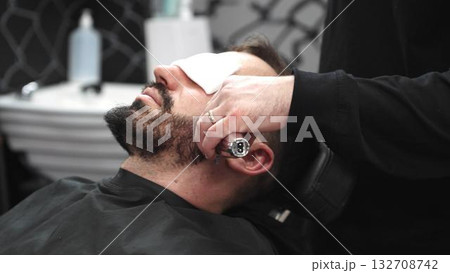 Professional barber grooming client's beard using electric razor, delivering precise trimming in contemporary barbershop setting 132708742