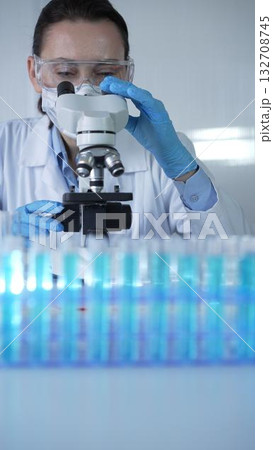 Female scientist wearing lab coat and protective blue gloves using microscope doing research in modern laboratory, portrait view. Low key lighting photo. Science and Medicine Female scientist wearing lab coat and protective blue gloves using microscope doing research in modern laboratory, portrait view. Low key lighting photo. Science and Medicine 132708745