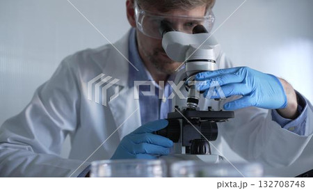 Male scientist researcher wearing blue gloves, protective glasses is using microscope in modern laboratory, portrait view. Low key lighting photo. Science and Medicine concepts Male scientist researcher wearing blue gloves, protective glasses is using microscope in modern laboratory, portrait view. Low key lighting photo. Science and Medicine concepts 132708748