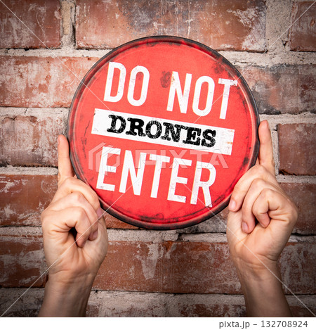 Round red warning sign with message do not enter DRONES, held by two hands against a brick wall 132708924