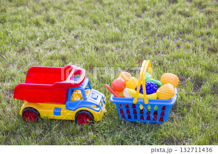 Car truckand a basket with toy fruit and vegetables. Bright plastic colorful toys for children outdoors on sunny summer day. Car truckand a basket with toy fruit and vegetables. Bright plastic colorful toys for children outdoors on sunny summer day. 132711436