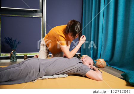 Masseuse making thai yoga massage. Woman therapist doing traditional massage treatment by elbow for male patient. Bald client lying on mat, meditating, enjoying hand massage in calm atmosphere. 132712043