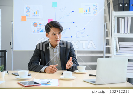 Young Asian businessman making FaceTime video calls, teleconference, having discussion and working on laptop computer sitting at the office, waving hand to webcam. Distant Communication Concept. Young Asian businessman making FaceTime video calls, teleconference, having discussion and working on laptop computer sitting at the office, waving hand to webcam. Distant Communication Concept. 132712235