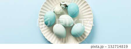 A collection of decorated blue Easter eggs sits on a white plate, showcasing various shades and patterns. The light background enhances the festive atmosphere of Easter, banner 132714819
