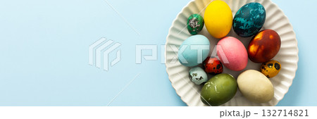 A variety of decorated Easter eggs in vibrant colors are creatively arranged on a white plate set against a soft blue background, symbolizing spring festivities and joy, banner 132714821