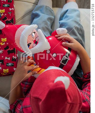 A young boy wearing a Santa hat sits on a sofa, holding an inflatable Santa toy. Cheerful holiday scene, perfect for Christmas, festive, and winter-themed projects. A young boy wearing a Santa hat sits on a sofa, holding an inflatable Santa toy. Cheerful holiday scene, perfect for Christmas, festive, and winter-themed projects. 132715718