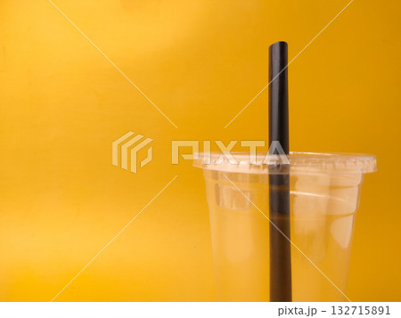The image shows a clear disposable plastic cup with a lid and a black straw on a yellow background. The image shows a clear disposable plastic cup with a lid and a black straw on a yellow background. 132715891