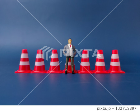 The image shows a small figurine of a man in a business suit standing among a line of red and white traffic cones. The image shows a small figurine of a man in a business suit standing among a line of red and white traffic cones. 132715897