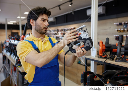 Worker man wearing overalls is holding a jigsaw tool in his hands Worker man wearing overalls is holding a jigsaw tool in his hands 132715921