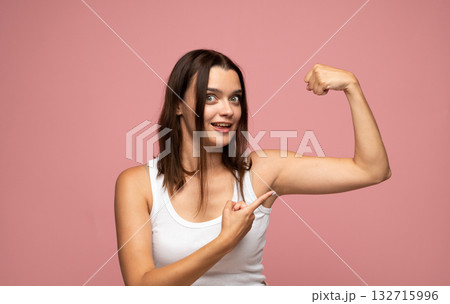 Excited young woman showing biceps and smiling proudly on pink background 132715996