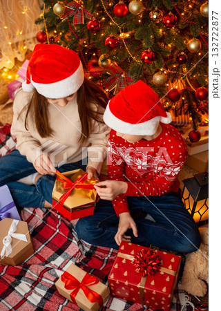 Horizontal photo of two children near the christmas tree, opening presents. Winter holidays, happy family atmosphere, festive interior. 132722877