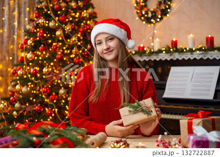 Happy young girl sitting near the table and holding packed and decorated present box. Gorgeous Christmas tree and piano on the background 132722887