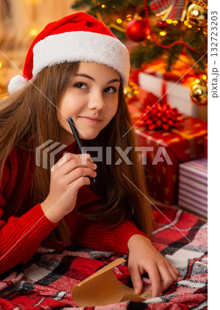 Teenage girl writing letter to Santa laying under Christmas tree. Happy small girl thinking off presents to mention in letter to Santa Teenage girl writing letter to Santa laying under Christmas tree. Happy small girl thinking off presents to mention in letter to Santa 132723203