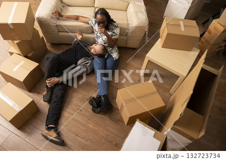 Multiethnic couple rests on floor surrounded by cardboard boxes during moving day. Caucasian boyfriend and black girlfriend bond while starting new chapter in cozy apartment after relocation. Multiethnic couple rests on floor surrounded by cardboard boxes during moving day. Caucasian boyfriend and black girlfriend bond while starting new chapter in cozy apartment after relocation. 132723734