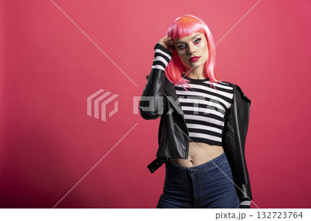 Pretty woman with pink bangs and bold eyeliner strikes confident pose in leather jacket and crop top during beauty campaign shoot. Glamorous lady casually modelling against isolated background. 132723764