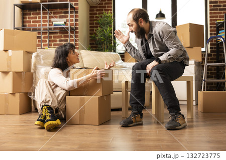 Young caucasian couple seated with cardboard boxes, having heated discussion on arranging items in new home. Angry wife on floor arguing with husband during moving day in brick wall apartment. Young caucasian couple seated with cardboard boxes, having heated discussion on arranging items in new home. Angry wife on floor arguing with husband during moving day in brick wall apartment. 132723775