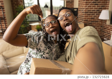 Playful black couple sticks tongues out at camera while holding house keys and cardboard box, enjoying fun moment in apartment. Excited spouses taking selfie with silly expressions in new home. Playful black couple sticks tongues out at camera while holding house keys and cardboard box, enjoying fun moment in apartment. Excited spouses taking selfie with silly expressions in new home. 132723828