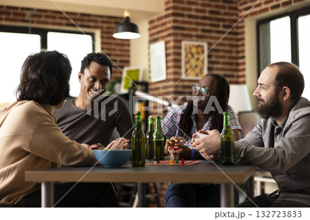 Small gathering of young adults engaged in conversation and playing indoor game around table. Diverse group takes break from board game, with everyone attentively listening to black man share a story. 132723833