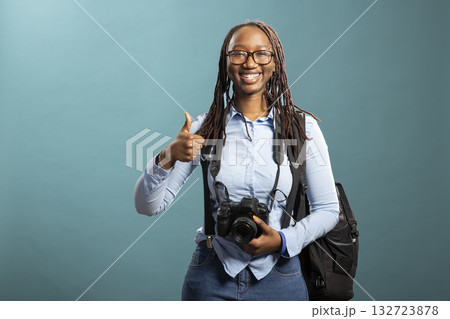 Energetic female photographer in studio with camera around her neck, giving thumbs up, excited for an upcoming photoshoot. African american woman doing an approval gesture against isolated background. Energetic female photographer in studio with camera around her neck, giving thumbs up, excited for an upcoming photoshoot. African american woman doing an approval gesture against isolated background. 132723878