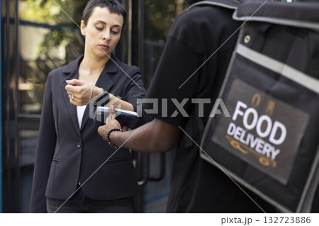 Takeaway courier waiting as a woman taps smart watch on a pos to pay for meal. Wireless transaction wraps up a seamless online order process for the food delivery, courier service. Takeaway courier waiting as a woman taps smart watch on a pos to pay for meal. Wireless transaction wraps up a seamless online order process for the food delivery, courier service. 132723886