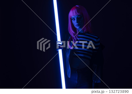 Stylish young woman with bright pink hair holds glowing blue neon tube in dark, cinematic studio. Female model wears a striped top and leather jacket, exuding cyberpunk attitude. Stylish young woman with bright pink hair holds glowing blue neon tube in dark, cinematic studio. Female model wears a striped top and leather jacket, exuding cyberpunk attitude. 132723890