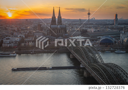 A breathtaking view of the Cologne skyline during sunset featuring the iconic Cologne Cathedral and the city's famous bridge. The warm hues of the evening sky create a stunning scene in Germany. 132728376