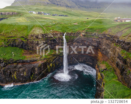 Dramatic Mulafossur waterfall plunging into the ocean with Gasadalur village and cliffs in the background. Dramatic Mulafossur waterfall plunging into the ocean with Gasadalur village and cliffs in the background. 132728654