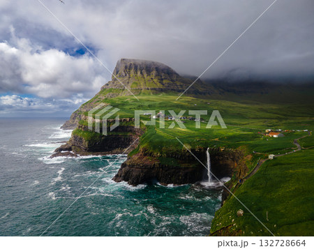 Dramatic Mulafossur waterfall plunging into the ocean with Gasadalur village and cliffs in the background. 132728664