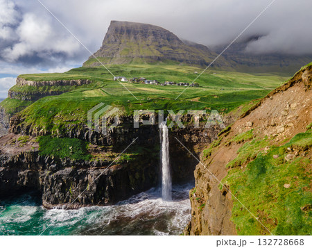 Dramatic Mulafossur waterfall plunging into the ocean with Gasadalur village and cliffs in the background. Dramatic Mulafossur waterfall plunging into the ocean with Gasadalur village and cliffs in the background. 132728668