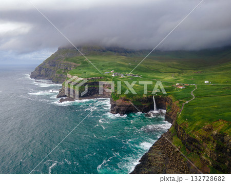 Dramatic Mulafossur waterfall plunging into the ocean with Gasadalur village and cliffs in the background. Dramatic Mulafossur waterfall plunging into the ocean with Gasadalur village and cliffs in the background. 132728682