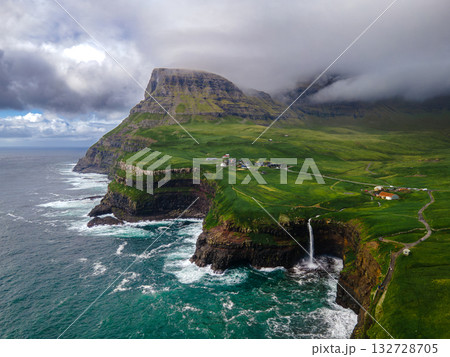 Dramatic Mulafossur waterfall plunging into the ocean with Gasadalur village and cliffs in the background. 132728705