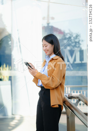 Confident Asian businesswoman smiling while using a smartphone outdoors, symbolizing technology, success, and modern professional lifestyle. Confident Asian businesswoman smiling while using a smartphone outdoors, symbolizing technology, success, and modern professional lifestyle. 132734116