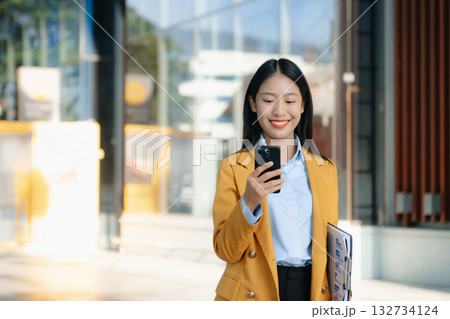Confident Asian businesswoman smiling while using a smartphone outdoors, symbolizing technology, success, and modern professional lifestyle. 132734124