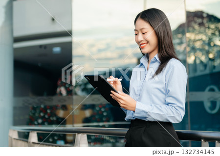 Asian woman with a smile standing holding notepad and tablet at out side office. big city on business center Asian woman with a smile standing holding notepad and tablet at out side office. big city on business center 132734145