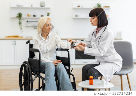 A female doctor measures the blood pressure of an elderly woman sitting in a wheelchair in a medical examination room. The patient is wearing casual clothes. A female doctor measures the blood pressure of an elderly woman sitting in a wheelchair in a medical examination room. The patient is wearing casual clothes. 132734234