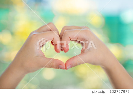 Two small hands forming a heart shape together with soft blurred natural background. Generation Alpha expressing love and following modern Gen Z social trends. 132737975