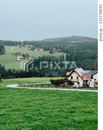 Green meadows and rolling hills create picturesque landscape, with winding road leading to traditional houses in Austrian countryside. Wooden farmhouses nestled among rolling alpine hills in Austrian 132739695