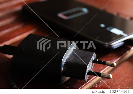 A black smartphone with a low battery icon is connected to a charger on a wooden surface. The charger is plugged into a power outlet. A black smartphone with a low battery icon is connected to a charger on a wooden surface. The charger is plugged into a power outlet. 132744162