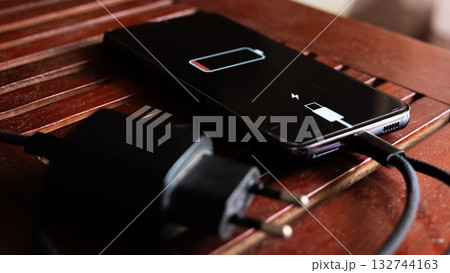 A black smartphone with a low battery icon is connected to a charger on a wooden surface. The charger is plugged into a power outlet. 132744163