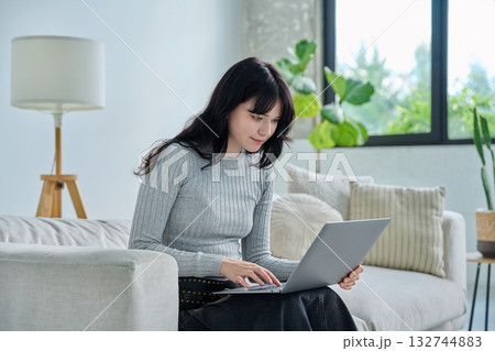 Young woman sitting on sofa typing on laptop, in home interior Young woman sitting on sofa typing on laptop, in home interior 132744883