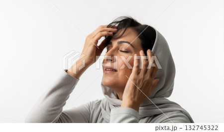 A middle-aged woman in a grey hijab with eyes closed touching her face and hair. Concept of stress relief, headache, or mindfulness. Isolated portrait on a white background A middle-aged woman in a grey hijab with eyes closed touching her face and hair. Concept of stress relief, headache, or mindfulness. Isolated portrait on a white background 132745322