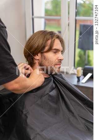 A hairdresser fastens a protective black cape on a male client before a haircut 132747104