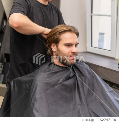 A hairdresser fastens a protective black cape on a male client before a haircut A hairdresser fastens a protective black cape on a male client before a haircut 132747110